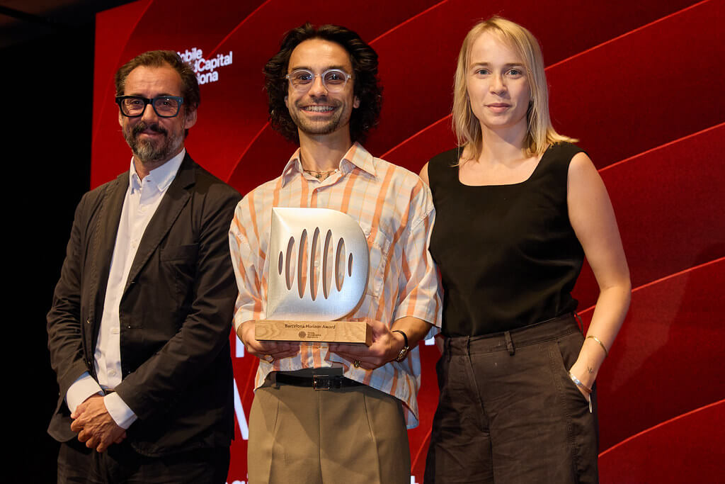Three people stand onstage; the person in the middle, smiling, holds a trophy. All wear business attire. Behind them is a red screen with white text. The mood is celebratory, suggesting an award event or ceremony.