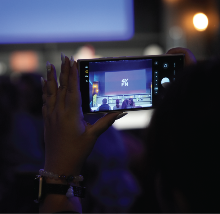 A person holds a phone horizontally, recording or photographing a large screen at an event. The phone display shows a stage with the logo “4YFN.” The background is dimly lit, with blurry figures and soft lighting.