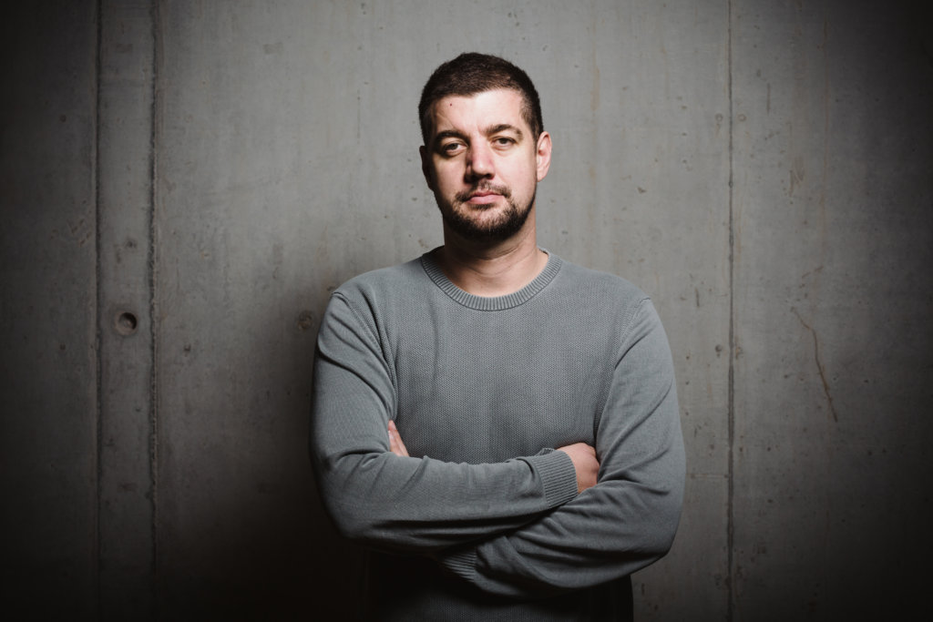 A man in a gray sweater stands with arms crossed, looking directly at the camera against a concrete wall background.