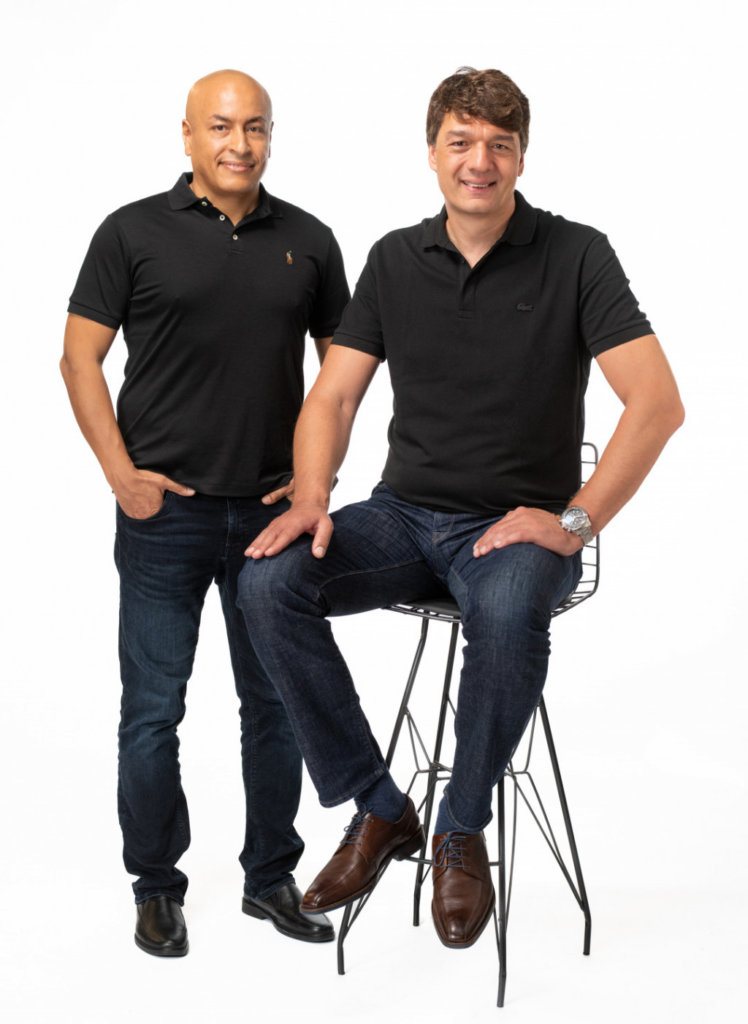 Two men posing against a white background. Both are wearing dark polo shirts and jeans. The man on the left is standing with his hand in his pocket, and the man on the right is sitting on a tall stool with his hands resting on his thighs.