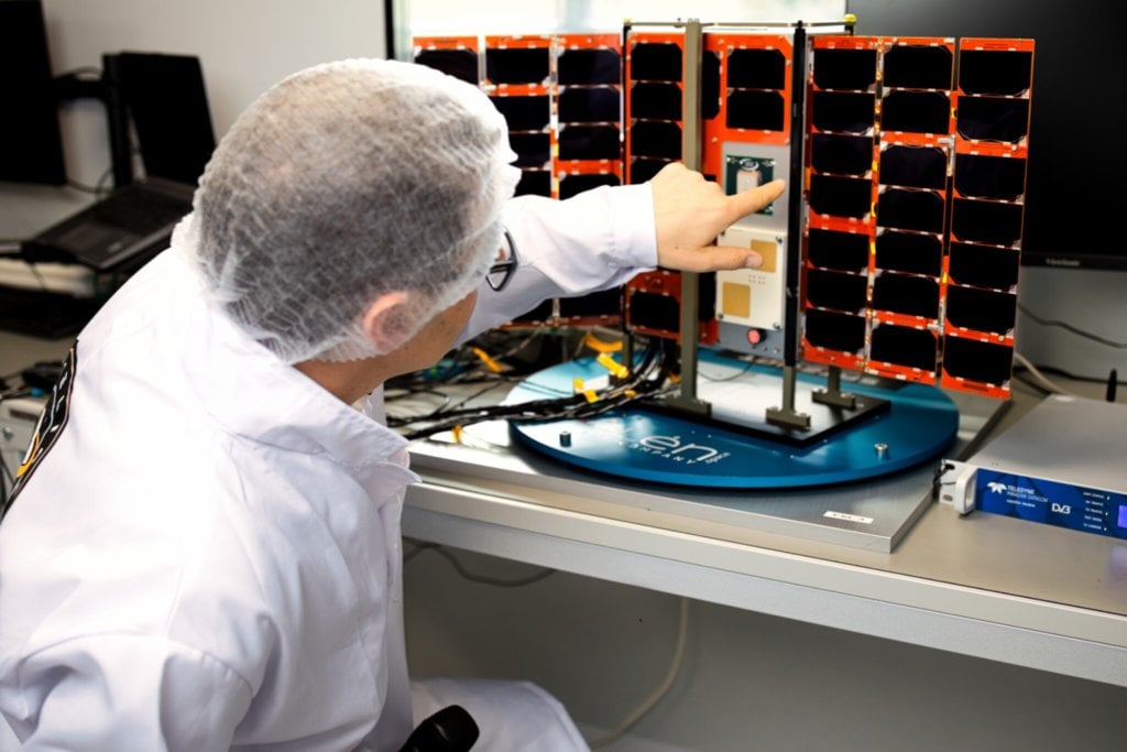 A person wearing a white lab coat, hair net, and glasses is pointing at a small satellite model with an array of solar panels in a lab setting. The individual is seated at a workbench with various electronic equipment.