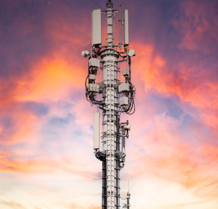 A tall cell tower stands against a vibrant sunset sky, featuring shades of pink, orange, and purple. The tower is equipped with multiple antennas and satellite dishes, capturing the technological elements against the natural beauty of the colorful, cloud-filled sky.