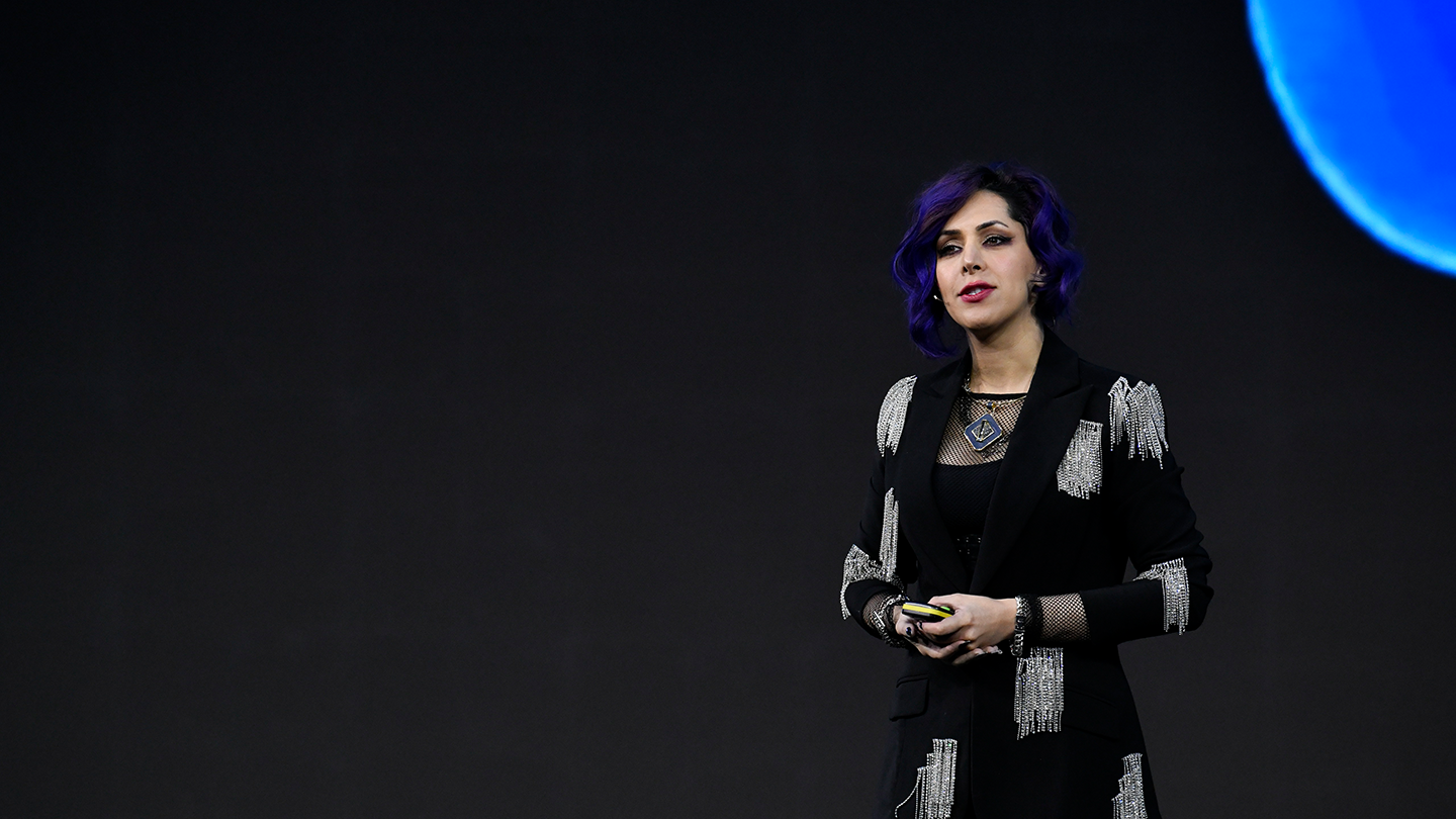 A person with purple hair stands on stage, wearing a black outfit embellished with silver details, exuding an aura as intriguing as someone asking, "What is GSMA Membership?" They hold a clicker and wear a necklace. The background features a dark screen with a partial view of a blue circular shape in the top right corner.