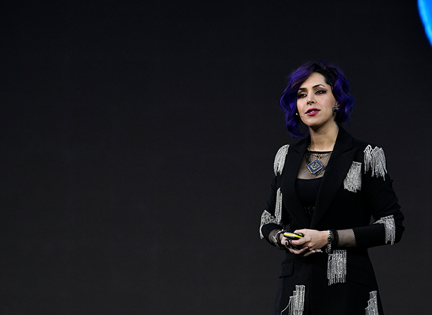 A person with wavy purple hair stands on stage against a dark background, wearing a black outfit with silver fringe details and holding a small object, confidently addressing an audience about What is GSMA Membership.