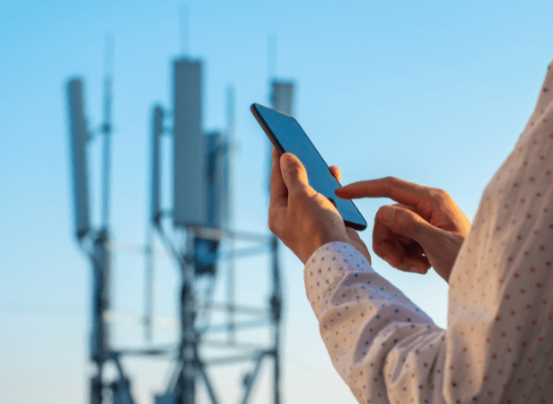 A person in a white, patterned shirt uses a smartphone outdoors. In the blurred background, several large cellular antennas and towers rise against a clear blue sky, hinting at MVNO services and a vibrant mobile network environment.