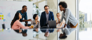 Six diverse professionals gather around a laptop in a bright meeting room. One woman stands and gestures to the laptop as the others, seated or standing, attentively watch and listen. Whiteboard with notes and sticky notes is visible in the background.