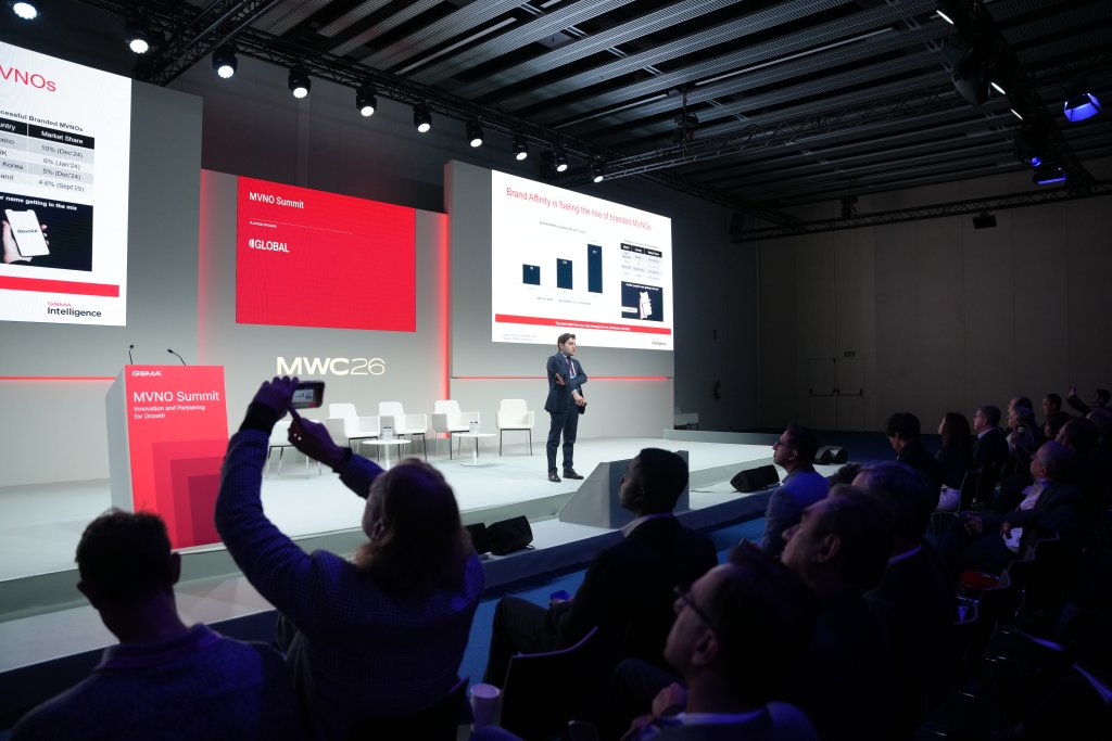 A man presents on stage at a tech conference with slides and an audience watching and taking photos.