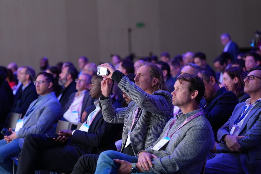 Audience seated at a conference, with one man taking a photo using his phone.
