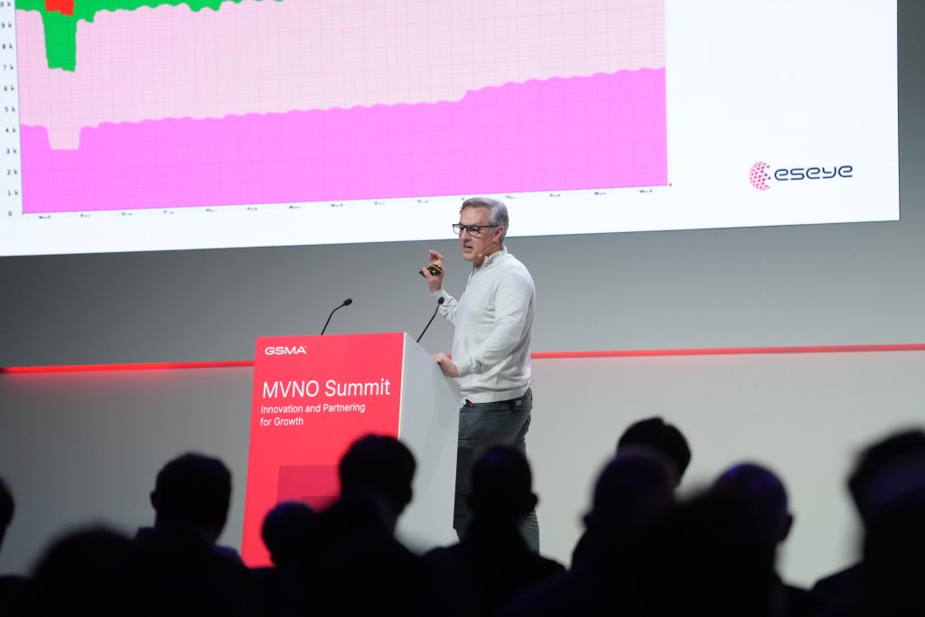 A man speaks at the MVNO Summit, presenting a graph on a large screen to an audience.