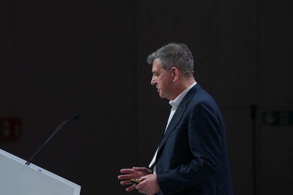 A man in a dark suit speaks at a podium with a microphone, gesturing with his hands.