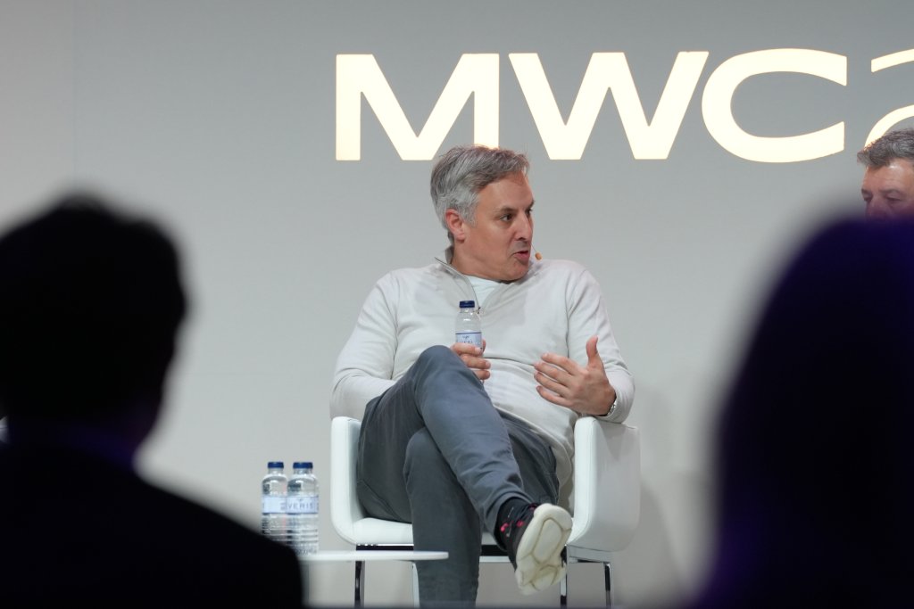 Man in a white sweater speaks while sitting on stage at MWC event, holding a water bottle.