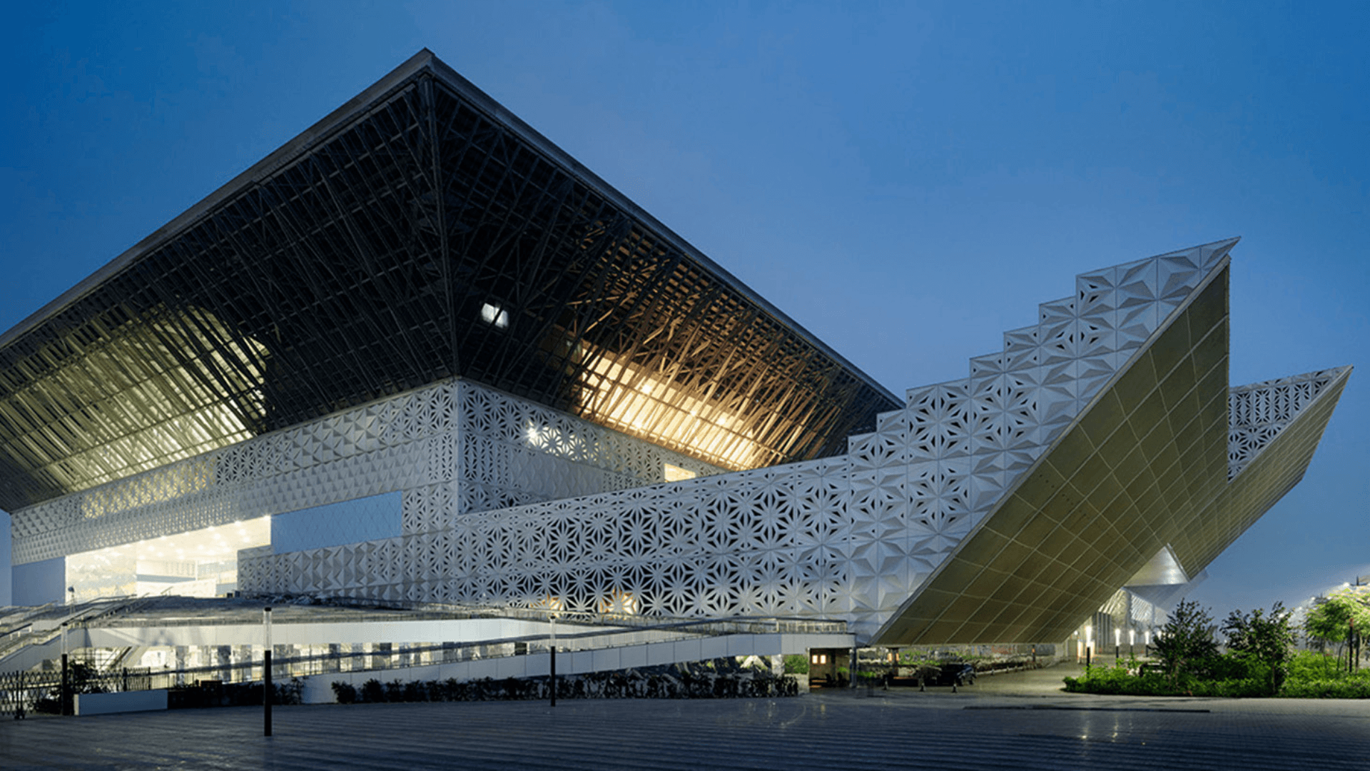 Modern building with geometric, angular design and patterned facade, illuminated in the evening with greenery nearby, serving as a Member gateway for innovation and collaboration through GSMA membership.