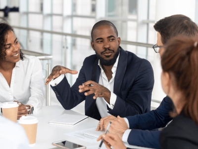 Four professionals sit around a white table in discussion. The man in the center, gesturing with his hands, appears to be leading a conversation about MVNO strategy. Two people take notes, and coffee cups are on the table. They are in a bright, modern office with large windows.