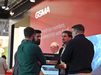Four men stand talking at a GSMA event booth, with a large red GSMA sign and tech screens in the background. Wearing business attire, they engage in conversation about MVNO strategies at this networking or conference setting, while other attendees are blurred behind them.