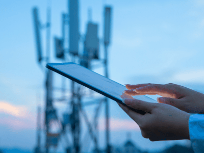 A person uses a tablet outdoors at dusk, with their hands touching the screen. In the blurred background, several large cellular towers with antennas rise into the sky, suggesting a focus on MVNO services or wireless communication infrastructure.