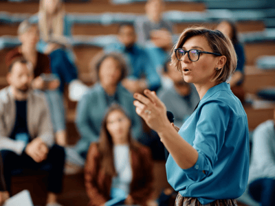 A person wearing glasses and a blue shirt is speaking energetically to a group of seated people in a lecture hall, delivering an engaging presentation about MVNOs. Focus is on the speaker, with the audience in the background, slightly blurred, listening attentively.
