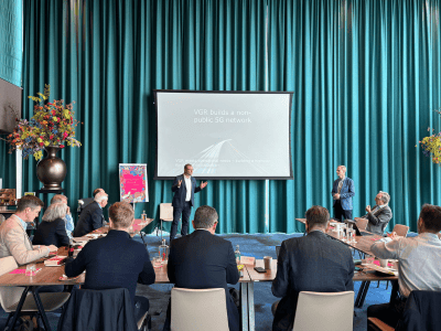 A man in a suit presents to an audience in a modern conference room with turquoise curtains. Another person stands nearby. Attendees sit at tables, listening and taking notes. A presentation on "VGR builds a non-public 5G network and explores MVNO partnerships" is displayed on a large screen.
