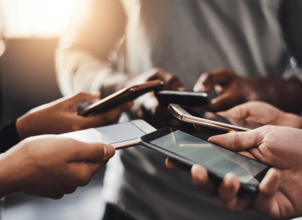 A group of people standing close together, each holding and using a smartphone. The photo focuses on their hands and the phones, with blurred backgrounds and warm lighting, suggesting social interaction or shared activity with technology.
