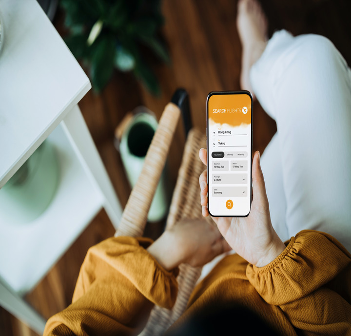 A person in white pants and a yellow long-sleeve top sits on a chair, holding a smartphone showing a “Search Flights” booking app. The surrounding area includes a small white table, a potted plant, and a green coffee cup on a wooden floor.