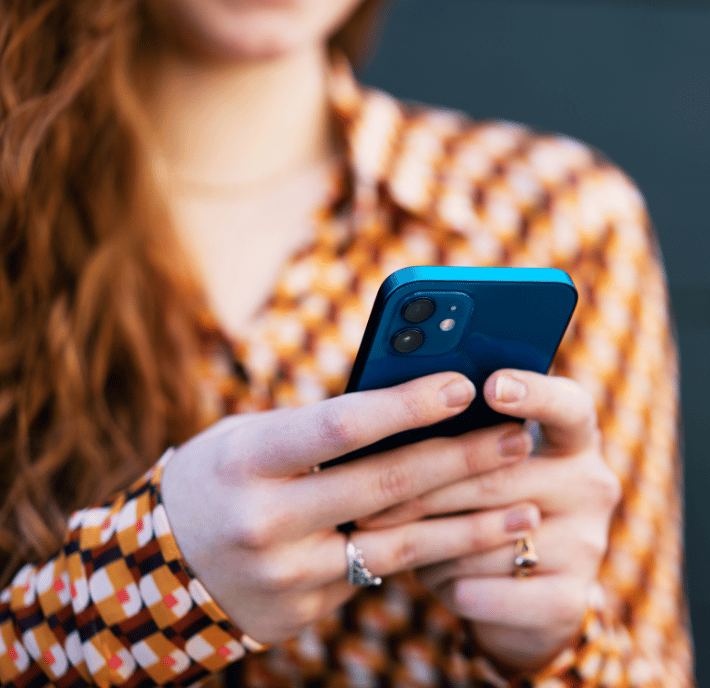 Woman with long red hair holding a blue smartphone and wearing a patterned shirt, background blurred.
