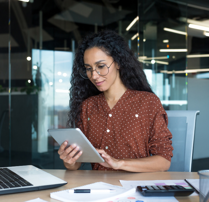 Woman with curly hair and glasses uses a tablet at her desk, researching GSMA Working Groups, with a laptop and office supplies nearby.