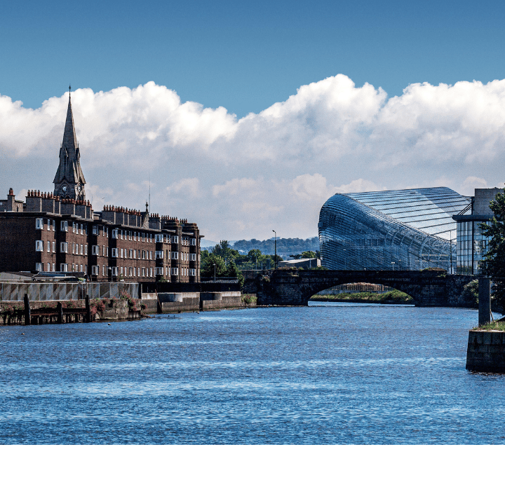 A river flows past old brick buildings, a church steeple, and a modern glass structure—much like the harmony found in GSMA Working Groups—under a clear blue sky.