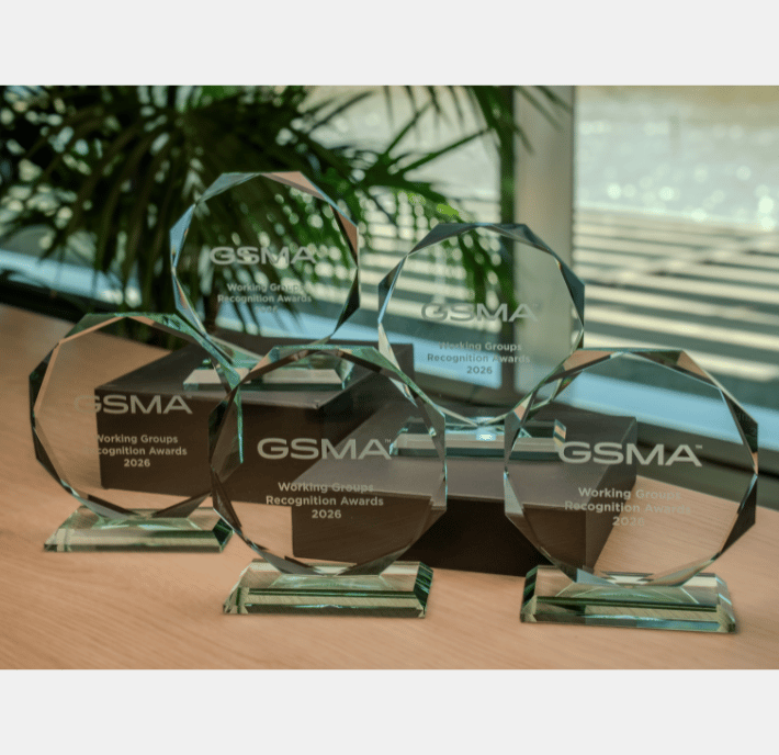 Five glass awards labeled "GSMA Working Groups Recognition Awards 2024" are displayed on a wooden surface, arranged in two rows. Sunlight streams through a window with leafy plants visible behind the Working Groups Recognition Awards winners.