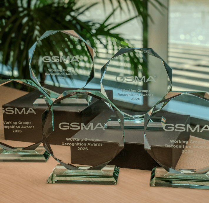 Five glass awards engraved with "GSMA Working Groups Recognition Awards 2026" are displayed on a wooden surface, with sunlight streaming through blinds and green leaves visible in the background, celebrating the achievements of GSMA Working Groups.