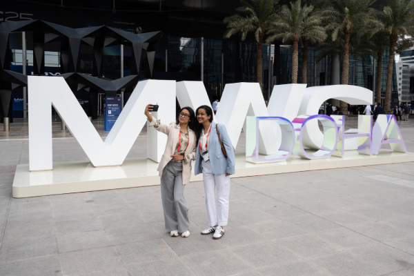 Two women stand smiling and taking a selfie in front of large, white "MWC DOHA" letters. Palm trees line the background, and a modern glass building is visible behind them. Both are dressed in business-casual attire and wearing event badges.