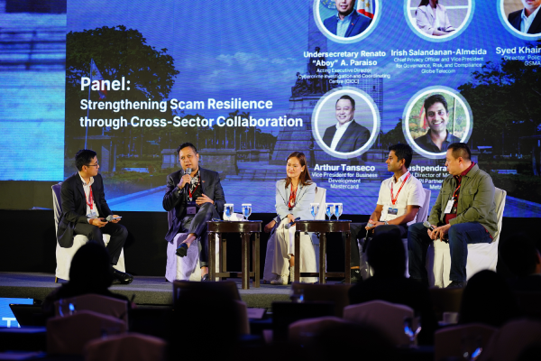 Five panelists, four men and one woman, sit on stage discussing scam resilience. Behind them, a screen displays the topic “Strengthening Scam Resilience through Cross-Sector Collaboration” with headshots, names, and titles. Audience members are visible in the foreground.