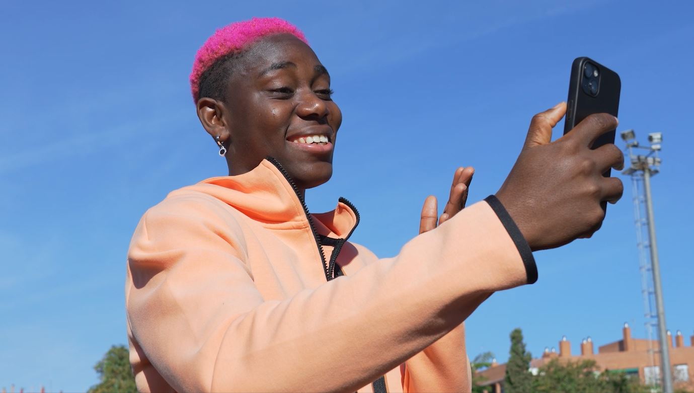 Asisat Oshoala with pink hair wearing a peach-colored jacket smiles and waves at a smartphone they are holding up for a video call. The background includes a clear blue sky and a few buildings and structures in the distance.