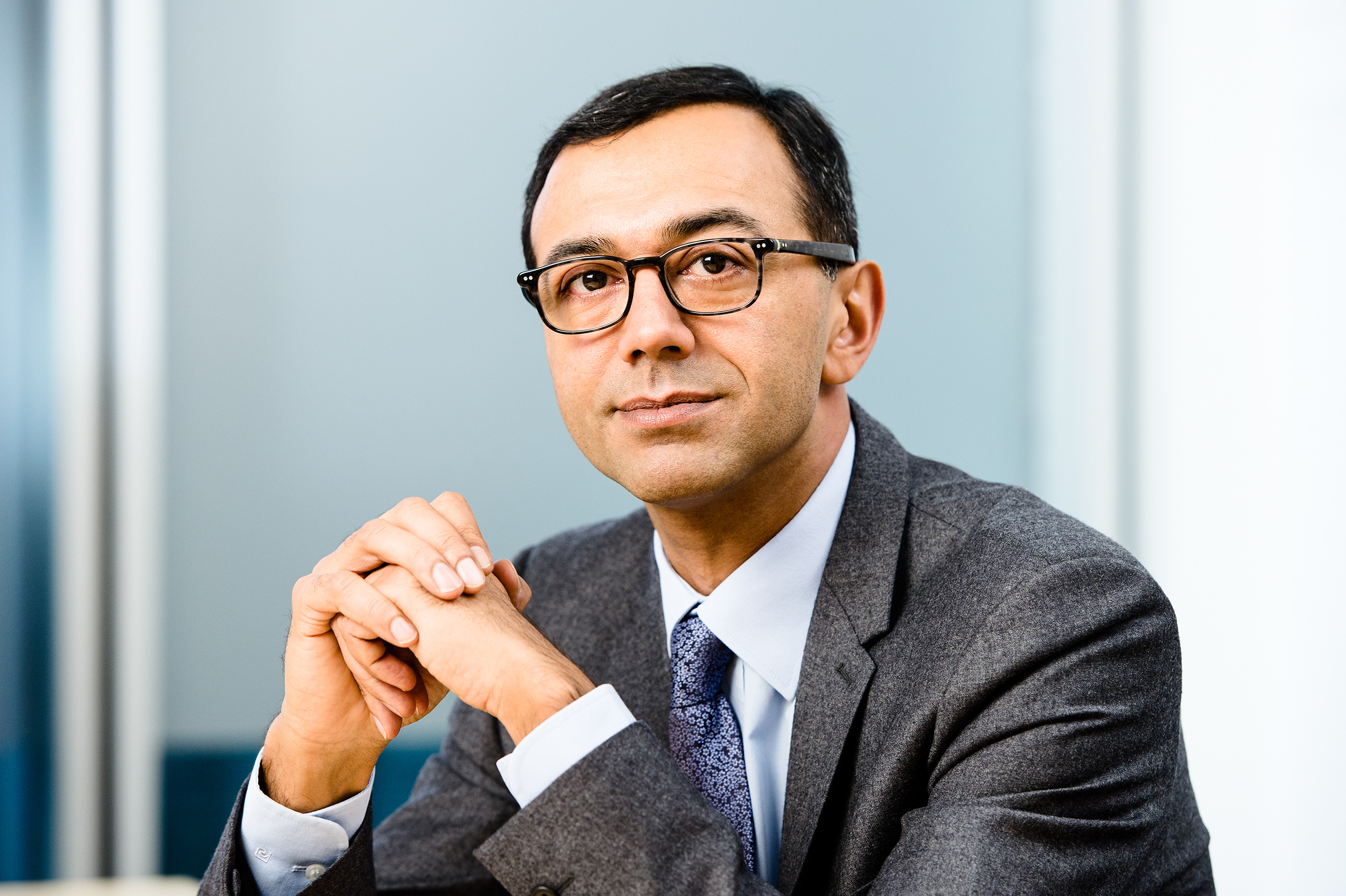 A man wearing glasses and a grey suit with a blue tie is sitting and looking directly at the camera. He has his hands folded under his chin against a backdrop with blue and grey tones.
