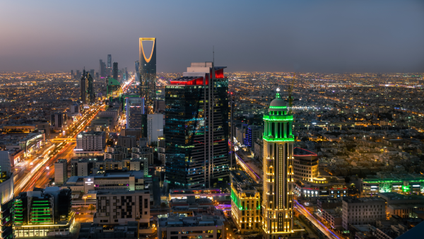 Aerial view of Riyadh at dusk, featuring modern skyscrapers with vibrant green and red lights, illuminated streets, and the iconic Kingdom Centre with a glowing arch, set against a sprawling cityscape under a fading blue sky.