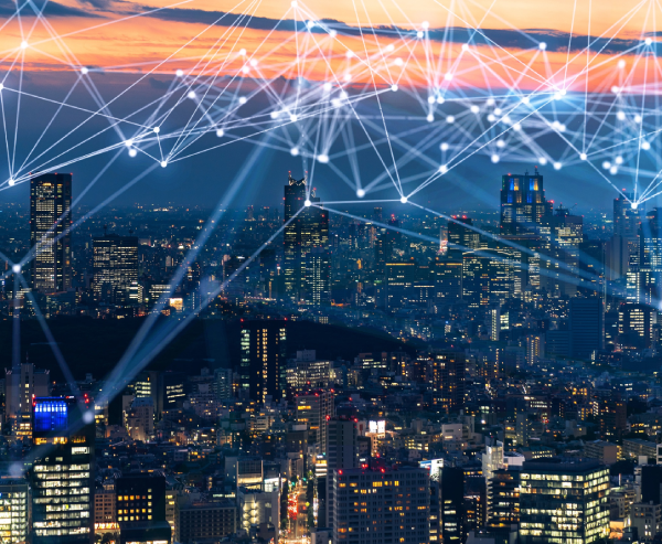 A city skyline at sunset with tall buildings illuminated by lights. Overlaying the city are glowing connected lines and nodes, symbolising digital connectivity or a network, stretching across the sky, representing technology and communication.