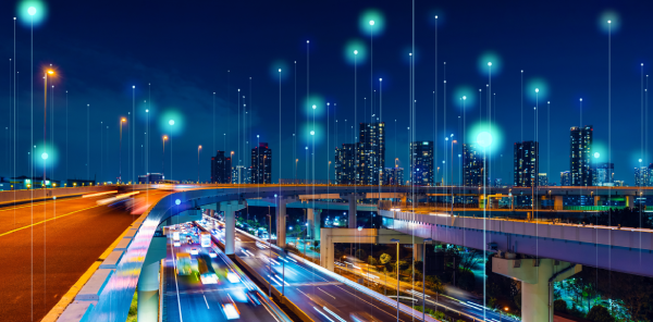 A nighttime cityscape shows tall buildings illuminated against the sky. Multi-level highways with blurred car lights suggest fast motion. Glowing digital dots and lines float above, symbolizing technology and connectivity in an urban setting.