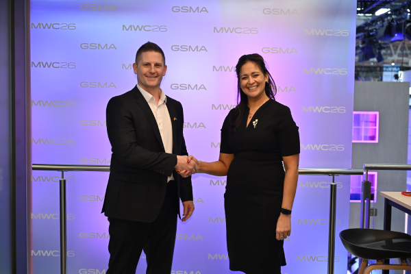 A man in a suit and a woman in a black dress shake hands and smile at a GSMA MWC26 event, standing in front of a branded backdrop with purple and blue lighting.