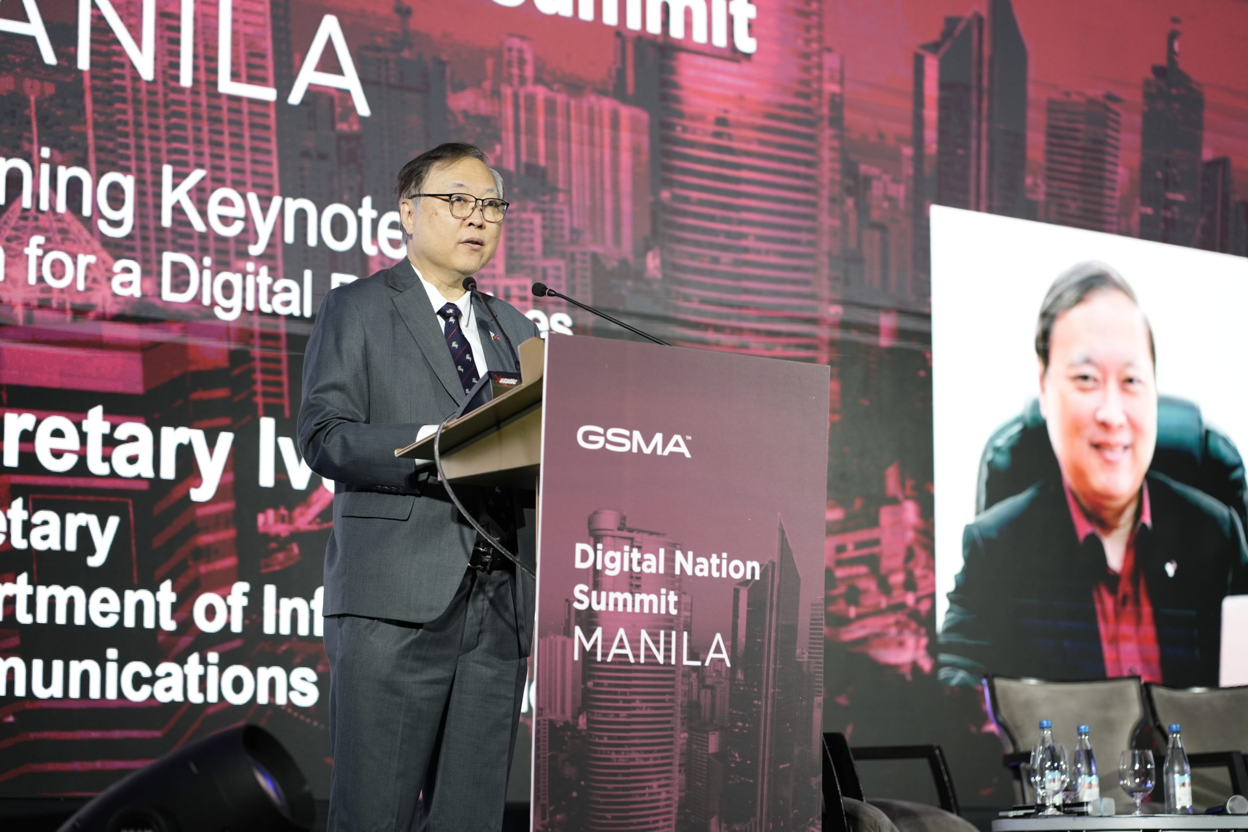 Secretary Ivan John E. Uy speaks at a podium with a microphone at the GSMA Digital Nation Summit in Manila. Behind him is a large screen displaying his photo and the event details in bold text.