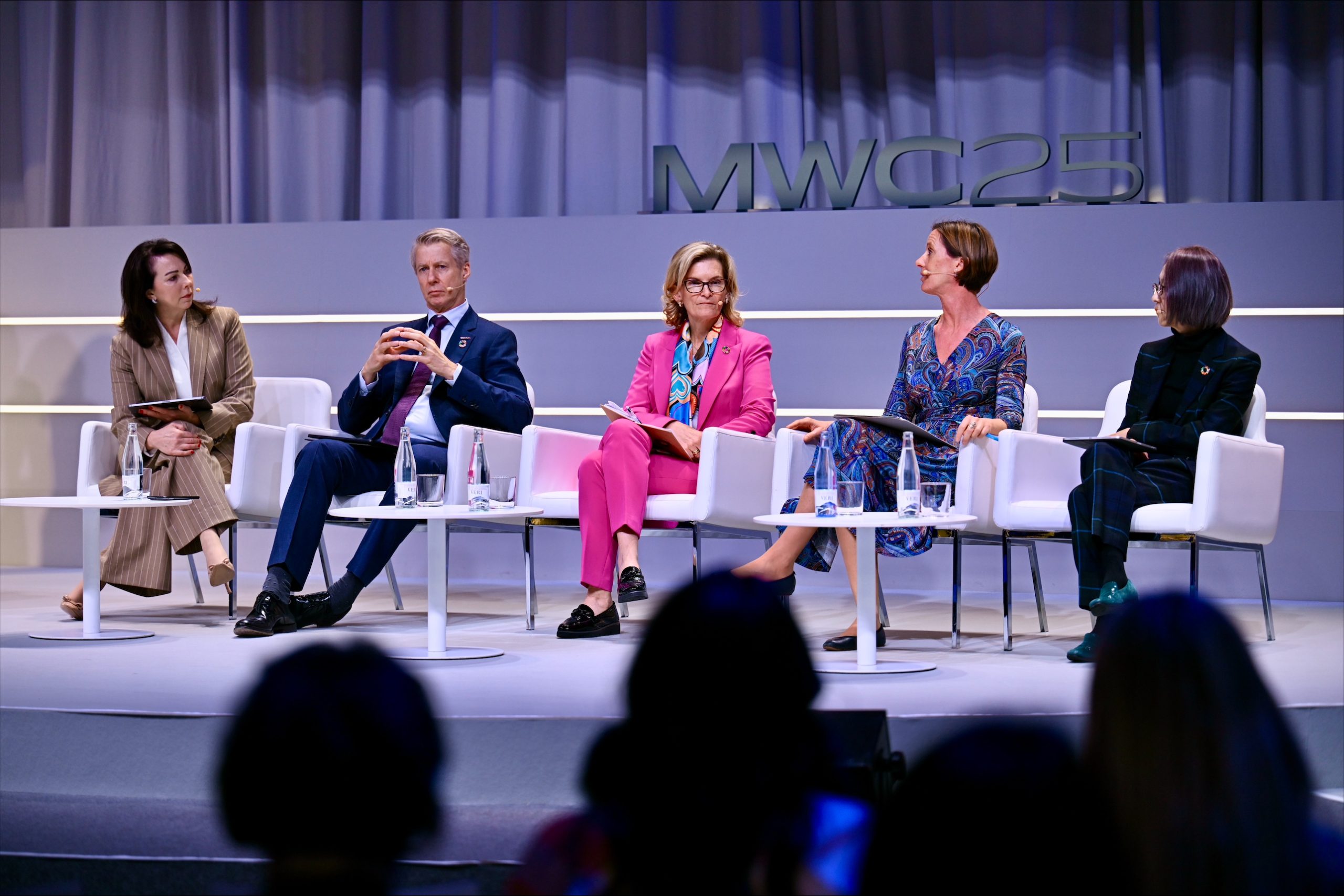 A panel of five individuals sitting on stage at MWC25. They are wearing formal business attire and are engaged in discussion. The background features a curtain with "MWC25" displayed. Each person holds a microphone or notepad, with glasses of water on the tables in front of them.