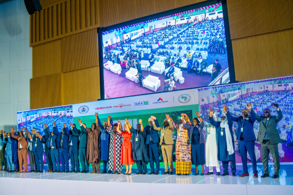 A large group of people in formal attire stand onstage, holding hands and raising them together. Behind them is a giant screen displaying a seated audience in a conference hall. A colourful banner decorates the stage backdrop.
