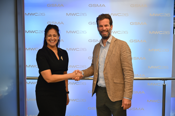 A woman in a black dress and a man in a tan blazer shake hands and smile at the camera. They stand in front of a blue backdrop with “GSMA” and “MWC26” logos repeated in gold. The setting appears formal and professional.