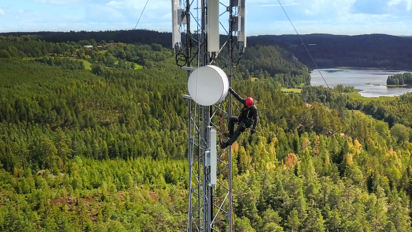 A worker wearing a red helmet and safety gear is working on a tall communication tower in a forested area. The tower is equipped with various antennae and dishes. The background features a lush green landscape with trees and a distant lake.