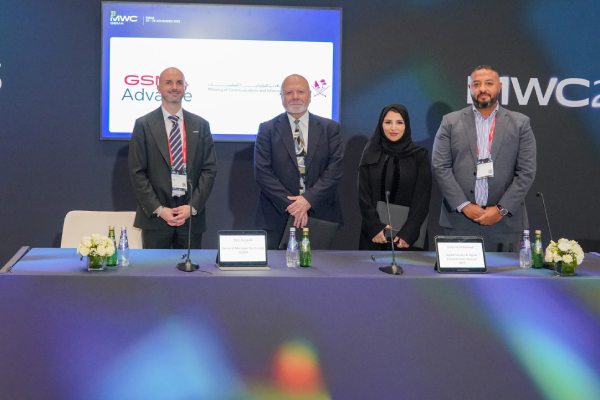 Four professionally dressed individuals stand behind a conference table with small flower arrangements and water bottles. A large screen behind them displays logos and text, including "GSMA Advancement" and Arabic writing. The setting appears formal and corporate.