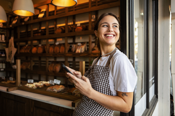 A smiling woman wearing a white T-shirt and a brown chequered apron leans against a window while holding a mobile phone. She is inside a bakery with wooden shelves displaying loaves of bread in the background.