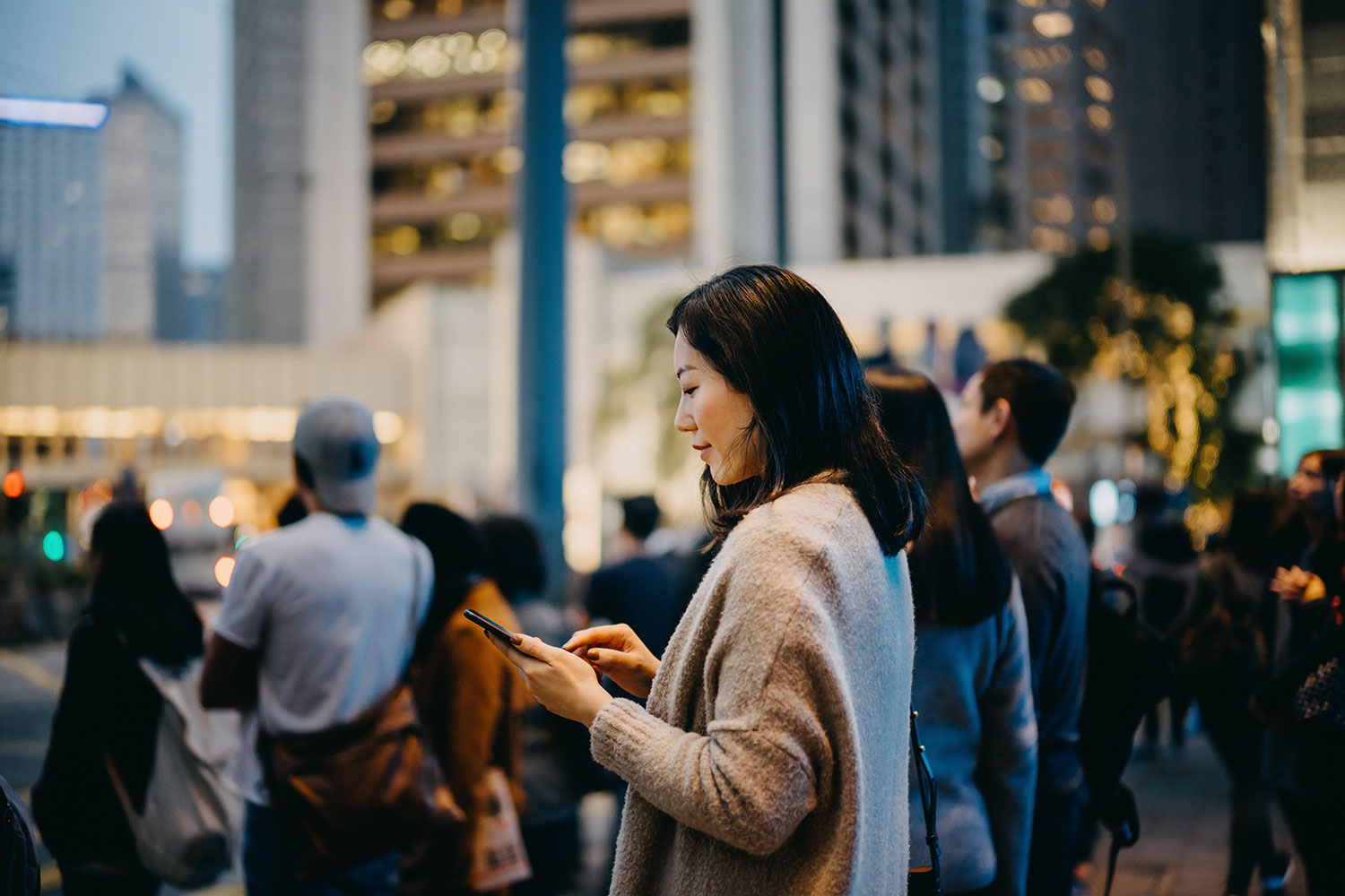 A woman is standing on a busy city street, focused on her phone. She is wearing a beige coat and has long dark hair. It's dusk, with city lights glowing softly in the background. People around her are also engaged with their phones or conversing, capturing an urban evening scene.