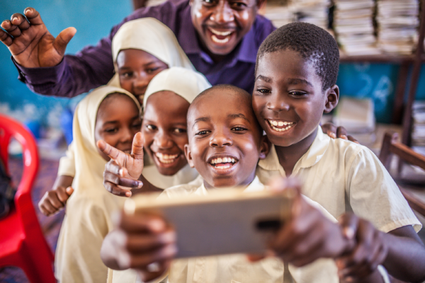 A group of smiling children in school uniforms and headscarves take a cheerful selfie with a smartphone, joined by an adult who is waving. They are indoors, surrounded by books and a red chair, creating a joyful, lively scene.