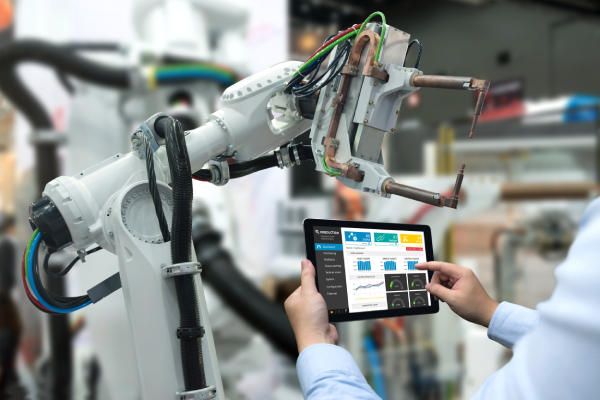 A person uses a tablet displaying analytics and data to control or monitor a large industrial robotic arm in a factory setting. The robotic arm has exposed wiring and metal components, indicating advanced automation technology.