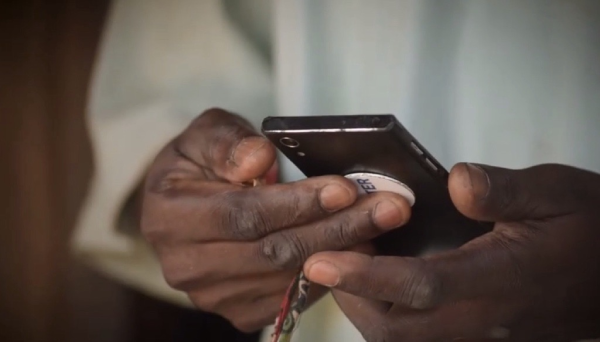 A close-up of two dark-skinned hands holding a black smartphone. The phone has a round white grip attached to the back, and a patterned lanyard is looped through it. The background is softly blurred, focusing attention on the hands and phone.