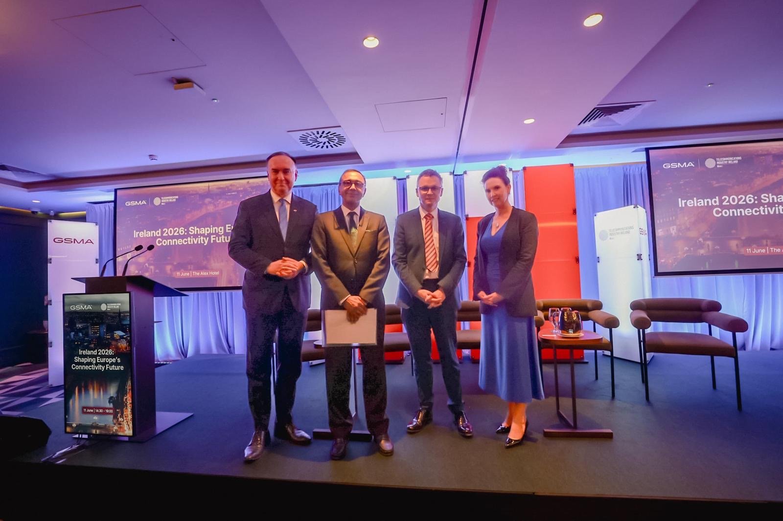 Four people in business attire stand side by side on a conference stage, smiling at the camera. Behind them are chairs, a table with water, and banners. A screen reads “Ireland 2026: Shaping Digital & Connectivity Futures.” GSMA