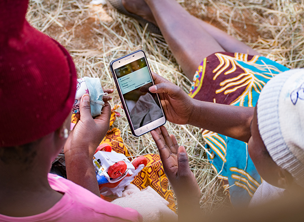 Two people are seated on the ground, one holding a baby. One person is holding a smartphone displaying a message. They are surrounded by straw or dried grass and are dressed in colorful clothing, with one person wearing a red hat and the other wearing a white hat.