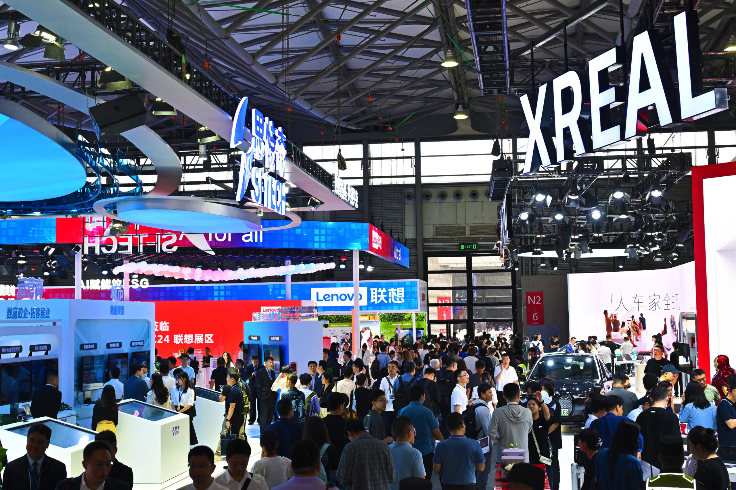 A busy tech expo with many people walking around and visiting various booths under large, lit-up signs, including "XREAL" and "Lenovo". The venue has a high ceiling with structural beams and multiple large displays showcasing products and technologies.